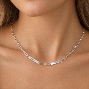 Può includere: Una collana a catena color argento con due piccole piastrine metalliche rettangolari integrate nelle maglie. Ogni piastrina è incisa con del testo, una riporta Merica e l'altra Jen, collegate da maglie di catena ovali allungate.