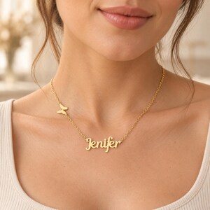 Può includere: Una collana color oro composta da una catenina sottile con un piccolo ciondolo a forma di farfalla posizionato lateralmente e una targhetta centrale con il nome Jenifer in corsivo, appoggiata sulla zona della clavicola.