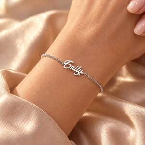 Può includere: Un bracciale a catena in metallo color argento con una targhetta centrale che riporta il nome Emily scritto in un carattere corsivo. Il bracciale è composto da delicati anelli circolari che si collegano all'elemento centrale su entrambi i lati.