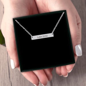 Può includere: Una collana a barra color argento con una catenina sottile e un ciondolo rettangolare inciso con il testo Luke 22:42. La collana è presentata all'interno di una scatola regalo quadrata nera, mostrando un design pulito e semplice.