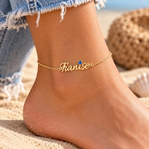 May include: A gold-toned anklet featuring a delicate chain and a custom nameplate with the script text "Franise" in a cursive font. The piece is accented with a single, small, round, deep blue gemstone set next to the nameplate.
