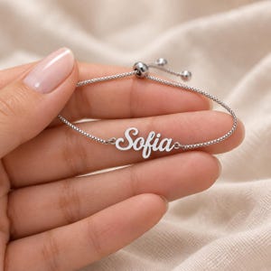 Può includere: Un braccialetto regolabile color argento con una sottile catena a maglie quadrate e una targhetta centrale con il nome Sofia scritto in corsivo. Il braccialetto include un meccanismo a perla scorrevole per regolare la lunghezza.