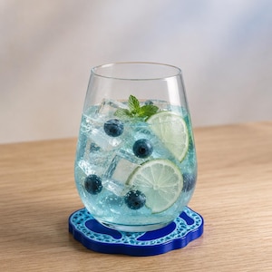 Può includere: Un bicchiere trasparente senza stelo riempito con un liquido azzurro, cubetti di ghiaccio, mirtilli freschi e due fette di lime, guarnito con un rametto di menta. Il bicchiere poggia su un sottobicchiere rotondo blu scuro con un motivo traforato.