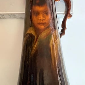 May include: A tall, dark brown ceramic pitcher with a glossy finish. The front of the pitcher features a detailed portrait of a person with dark hair and a collared shirt. It has a curved handle on the right side and a wide, flared pouring spout.