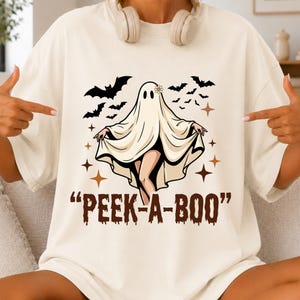 May include: A cream-colored t-shirt featuring a graphic of a ghost wearing a small flower on its head, surrounded by black bats and brown four-pointed stars. The text "PEEK-A-BOO" is printed in a brown, dripping font below the ghost illustration.