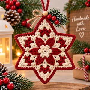 May include: A star-shaped crochet ornament featuring a central cream-coloured flower surrounded by deep red and cream petals. The ornament is topped with a small wooden bead, a jute twine bow, and a sprig of green pine needles with red berries.