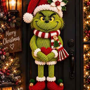 Könnte beinhalten: Ein gehäkelter Wandbehang in Form einer grünen Kreatur mit gelben Augen, die eine rot-weiße Weihnachtsmütze, einen rot-weiß gestreiften Schal mit einem kleinen Baum-Anhänger und rote Elfenstiefel mit goldenen Glöckchen trägt.