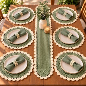 May include: A dining table setting featuring six round sage green crocheted placemats with cream scalloped edges, a matching long table runner, and six white plates topped with sage green napkins held by cream-colored beaded napkin rings.