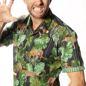 May include: A man wearing a short-sleeved button-down shirt with a jungle print featuring green leaves and leopard spots. He is wearing blue jeans and has a serious expression on his face.