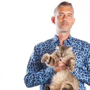 Puede incluir: Un hombre con una camisa azul con estampado de leopardo est&aacute; sosteniendo un gato marr&oacute;n y gris. El gato est&aacute; mirando a la c&aacute;mara con una expresi&oacute;n seria.