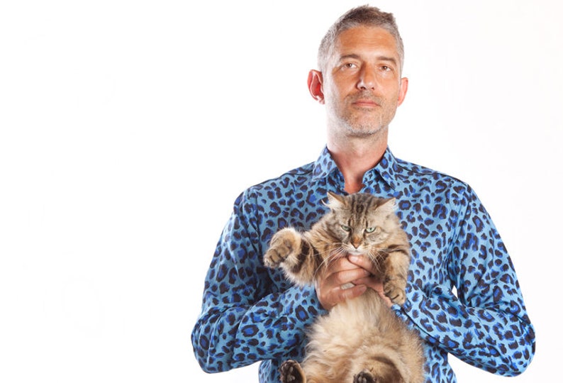 Puede incluir: Un hombre con una camisa azul con estampado de leopardo est&aacute; sosteniendo un gato marr&oacute;n y gris. El gato est&aacute; mirando a la c&aacute;mara con una expresi&oacute;n seria.