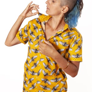 May include: A yellow short-sleeved button-down shirt with a black and white beetle print. The shirt is unbuttoned and the person is adjusting the collar.