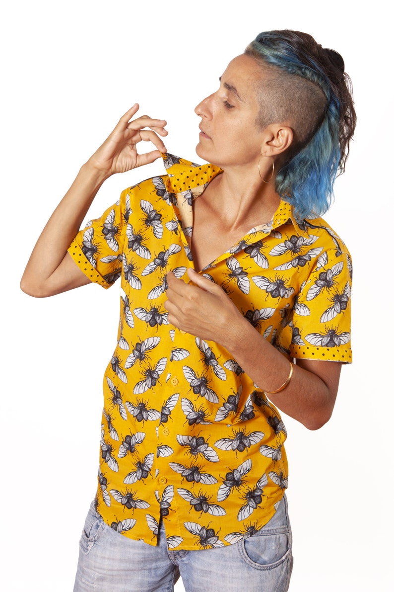 May include: A yellow short-sleeved button-down shirt with a black and white beetle print. The shirt is unbuttoned and the person is adjusting the collar.