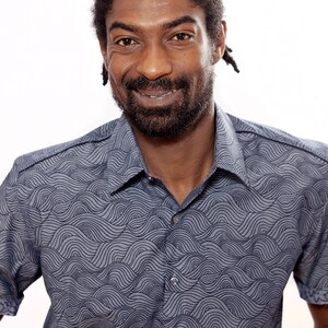 Pu&ograve; includere: Un uomo con la barba e dreadlocks corti indossa una camicia blu a maniche corte con un motivo a onde.