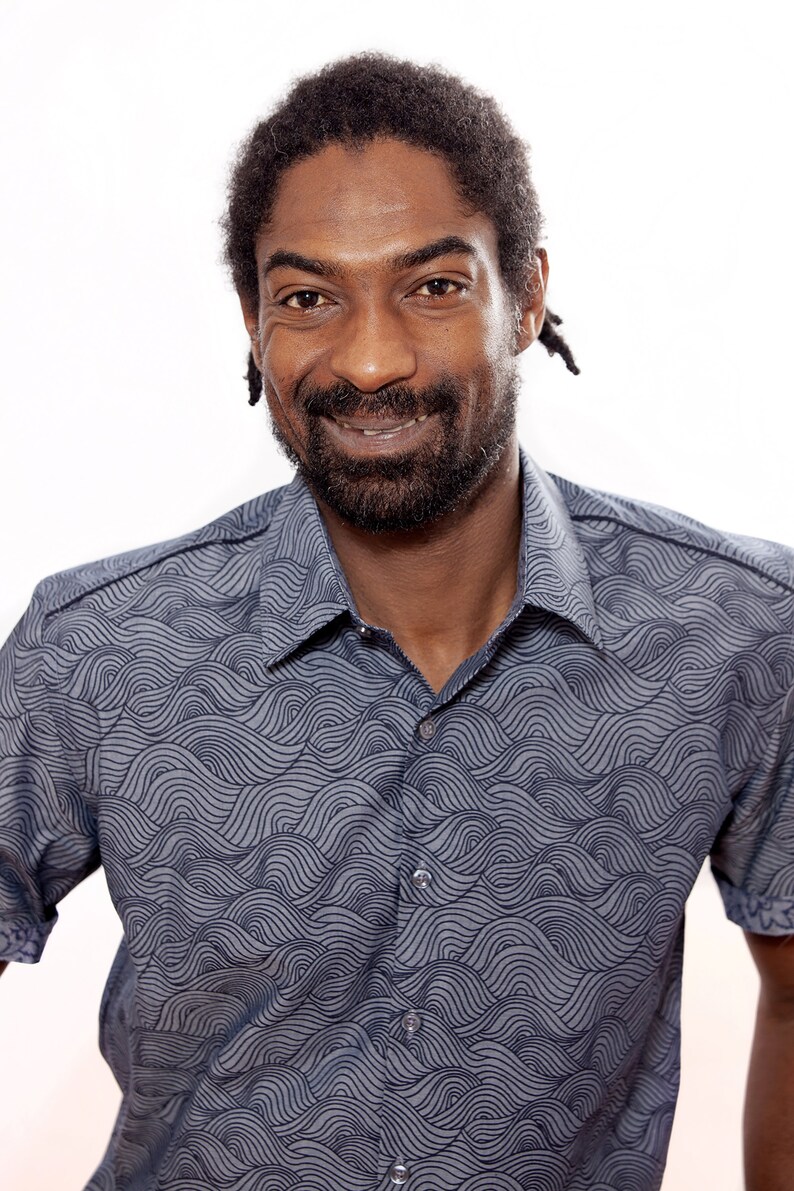 Pu&ograve; includere: Un uomo con la barba e dreadlocks corti indossa una camicia blu a maniche corte con un motivo a onde.