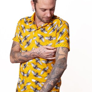 May include: A yellow short-sleeved button-down shirt with a black and white insect print. The shirt has a collar and a button closure. The person wearing the shirt has tattoos on both arms.