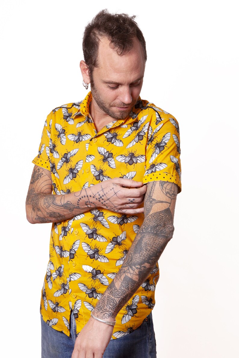 May include: A yellow short-sleeved button-down shirt with a black and white insect print. The shirt has a collar and a button closure. The person wearing the shirt has tattoos on both arms.