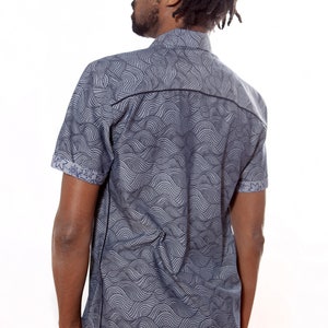 Pu&ograve; includere: Un uomo indossa una camicia blu a maniche corte con un motivo a onde. La camicia ha un bordo nero intorno al colletto e ai polsini.