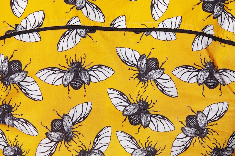 May include: Yellow fabric with a repeating pattern of black and white beetle illustrations. The beetles have wings and are drawn in a vintage style.