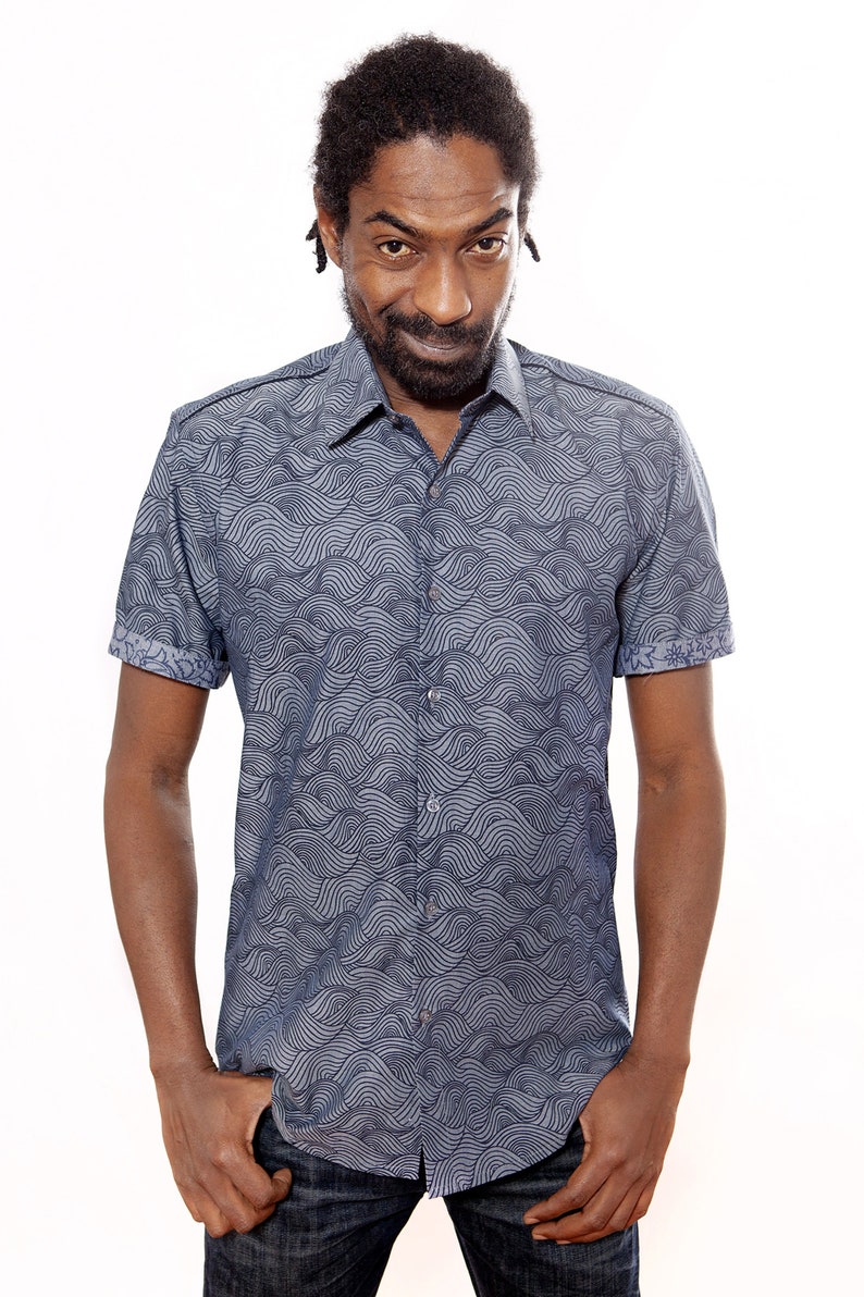Pu&ograve; includere: Un uomo indossa una camicia blu a maniche corte con un motivo ondulato. La camicia ha una chiusura con bottoni sul davanti e un colletto.