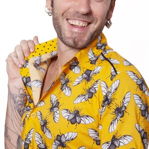 May include: A yellow short-sleeved shirt with a black and white beetle print. The shirt has a button-down collar and a black trim around the collar and sleeves.