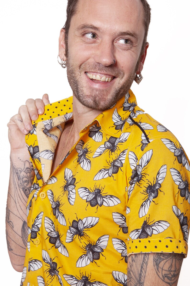 May include: A yellow short-sleeved shirt with a black and white beetle print. The shirt has a button-down collar and a black trim around the collar and sleeves.