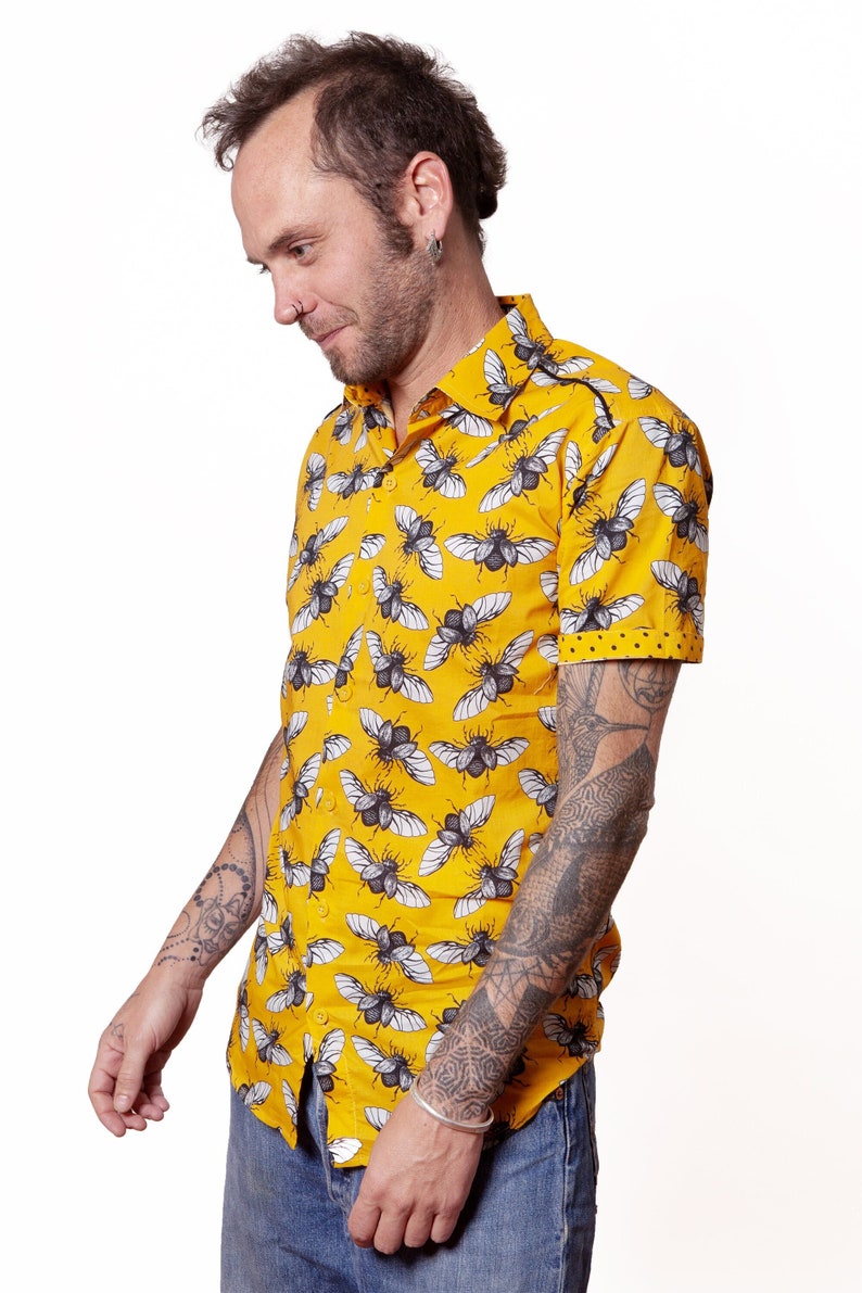 May include: A yellow short-sleeved button-down shirt with a black and white beetle print. The shirt has a collar and a pocket on the left chest. The model is wearing blue jeans and has tattoos on their arms.