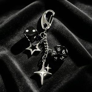 May include: A silver-toned metal keychain featuring a lobster clasp, a small black six-sided die with white pips, a black twenty-sided die with white numbers, a silver open-star charm, and a four-pointed silver star charm with a smaller star accent.