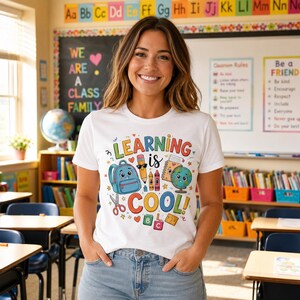 Peut inclure: Un t-shirt blanc à manches courtes avec une impression graphique colorée portant le texte "LEARNING is COOL!" entouré d'illustrations d'un sac à dos bleu, d'un crayon avec des lunettes, d'un globe, de ciseaux, de crayons et de blocs alphabétiques.