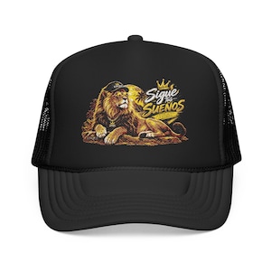 May include: A black trucker hat featuring a graphic print of a lion wearing a cap and a chain. The lion is lying down in front of a large yellow sun. The text reads Sigue Tus Sueños with a crown icon above the words in a stylised yellow font.