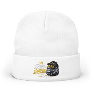 May include: A white cuffed beanie featuring a graphic print of a lion wearing a black cap and gold sunglasses. The text "Sigue tus sueños" is printed in gold and white lettering next to the lion, with the word "ENQUI" printed below the lion.