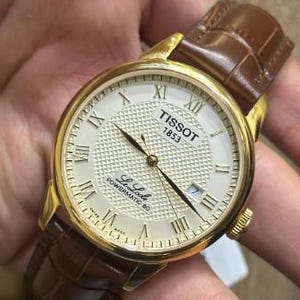 May include: A Tissot 1853 Le Locle Powermatic 80 wristwatch with a gold-tone round case, a white textured dial featuring Roman numeral hour markers, and a brown leather strap with a crocodile-embossed pattern and a gold-tone buckle closure.