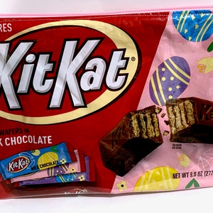 May include: A pink zippered pouch with a Kit Kat chocolate bar design. The pouch features a red and yellow Kit Kat logo, a broken Kit Kat bar, and a pink background with Easter eggs. The text "Kit Kat", "SP WAFERS IN MILK CHOCOLATE", and "NET WT 9.5 OZ (272 g)" is visible on the pouch.