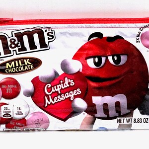 May include: A white zippered pouch with a red zipper pull. The pouch is decorated with a red M&M's character holding a red heart with the text "Cupid's Messages". The pouch also features a variety of M&M's candies with Valentine's Day messages, including "Be Mine" and "I <3 U". The text "3X the Chocolate" and "NET WT 8.83 OZ (250." is also visible on the pouch.
