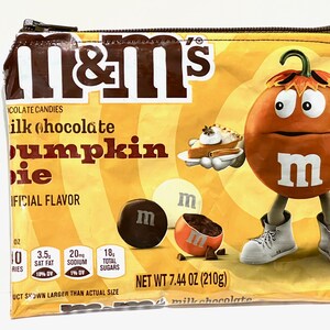 May include: A yellow and orange M&M's candy pouch with a pumpkin-shaped M&M character holding a slice of pumpkin pie. The pouch is labeled "Milk Chocolate Pumpkin Pie" and has a net weight of 7.44 oz (210g).