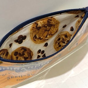 May include: Clear, rectangular pencil case with a blue zipper. The interior lining features a pattern of chocolate chip cookies and chocolate chips on a white background. The exterior shows the Ghirardelli logo and text.