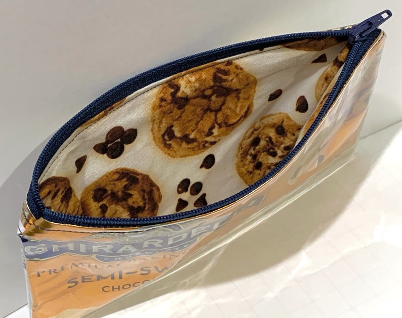 May include: Clear, rectangular pencil case with a blue zipper. The interior lining features a pattern of chocolate chip cookies and chocolate chips on a white background. The exterior shows the Ghirardelli logo and text.