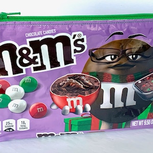 May include: A purple and green zippered pouch with a festive holiday design featuring the M&M's logo, a chocolate M&M character wearing a scarf and glasses, and a chocolate brownie with red and green M&M's. The pouch is labeled "Chocolate Candies" and "Net Wt 9.50 oz (269.3g)".