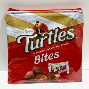 May include: Red zippered pouch with a graphic of a cartoon turtle wearing a top hat and bow tie. The pouch has the text "DeMet's Turtles The Original Caramel Nut Cluster Bites" and a smaller image of a candy bar with the text "Turtles Bites".