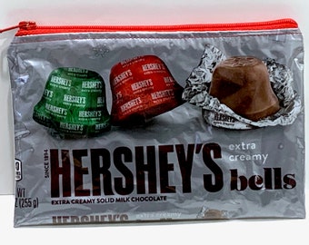 May include: A clear plastic zippered pouch with a gray background featuring Hershey's Extra Creamy Bells chocolate. The pouch has a red zipper and shows three chocolate bells in their foil wrappers. The text on the pouch reads "Hershey's Extra Creamy Bells, Since 1894, WT 2 (255 g), Extra Creamy Solid Milk Chocolate."