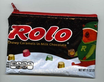 May include: A red and black zippered pouch with the Rolo logo and a cartoon bear holding a Rolo candy. The pouch is made of a shiny, plastic-like material and has a white background with a snowy scene. The text on the pouch reads "Chewy Caramels in Milk Chocolate" and "Net Wt 11 oz (31".