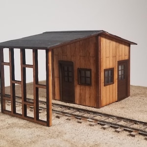 May include: A miniature wooden train depot model with a dark roof, brown walls, and a partial open-air structure. The model includes doors, windows, and sits on a sandy surface with miniature train tracks.