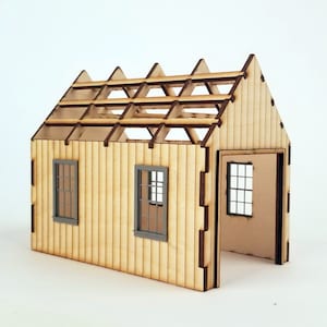 May include: A miniature wooden house model with an open door and two windows. The unfinished roof structure is visible. The walls are made of light-colored wood with vertical grooves, and the window frames are gray.