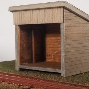 May include: A small wooden model of a train shelter with a brown interior and a light brown exterior. The shelter has a simple design with a pitched roof and a single door.