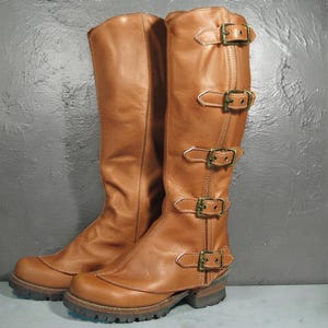 Könnte beinhalten: Ein Paar kniehohe Stiefel aus hellbraunem Leder mit fünf verstellbaren Riemen und antikfarbenen Messingschnallen, die vertikal an der Außenseite verlaufen. Die Stiefel haben eine abgerundete Zehenpartie, eine dicke Profilsohle und einen Blockabsatz.