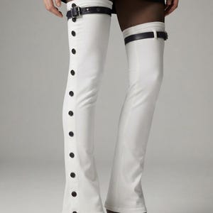 May include: White over-the-knee leg warmers featuring a vertical row of black buttons along the outer side of each leg. Each warmer is secured at the top with a dark brown leather-like strap and a metal buckle, paired with dark brown heels.
