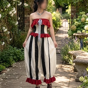 Könnte beinhalten: Ein trägerloser Jumpsuit mit einem Oberteil aus vertikalen schwarzen und weißen Streifen, akzentuiert durch rote Rüschen am Ausschnitt und an der Taille. Die weite Hose hat schwarze und weiße Streifen, einen roten Rüschensaum und ein hellblaues Band.
