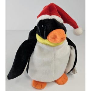 May include: A plush penguin toy featuring black and white fur, a bright orange beak, and orange feet. The penguin has a yellow band around its neck and is wearing a red and white fabric Santa hat that hangs down to the side of its head.
