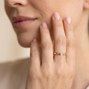 May include: A gold-toned open band ring featuring two small, flat, circular ends. Each end is engraved with a single uppercase letter, specifically the letter M on the left side and the letter J on the right side of the open gap.