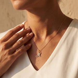 May include: A gold-toned necklace featuring a delicate chain and a single pendant shaped like the cursive letter M. The pendant hangs centrally from the chain, which rests against the collarbone area. The metal has a polished, reflective finish.